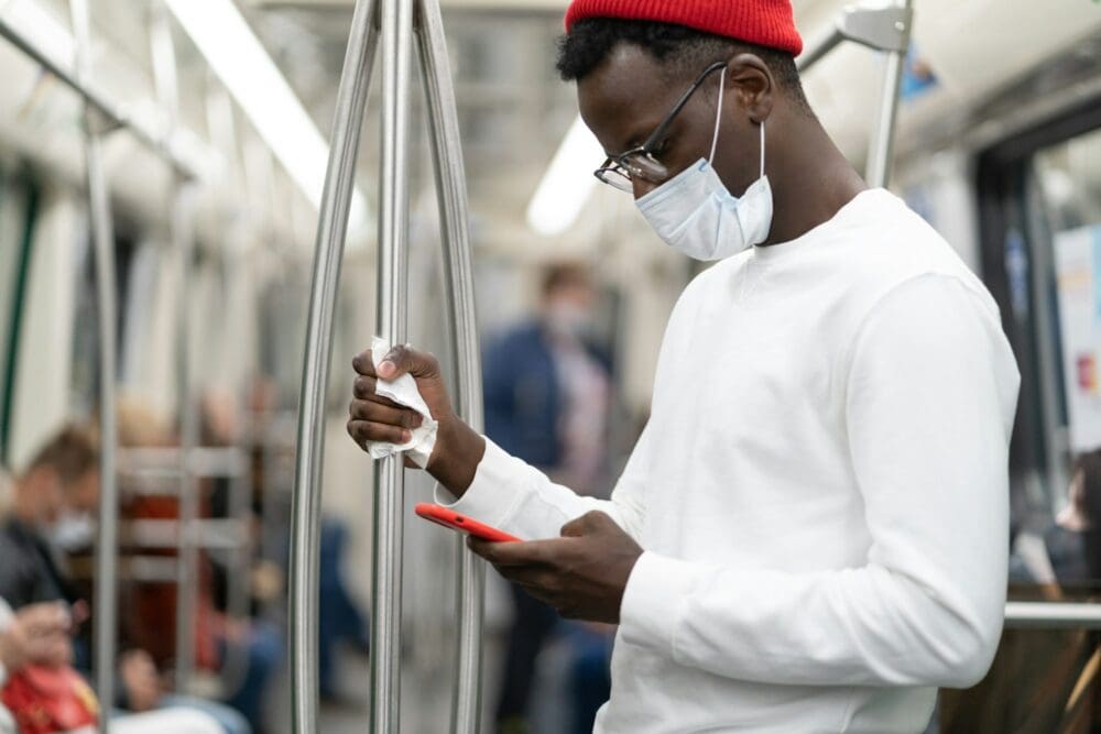 Black millennial man wearing face mask, using mobile phone, holding handrail in train. Covid-19