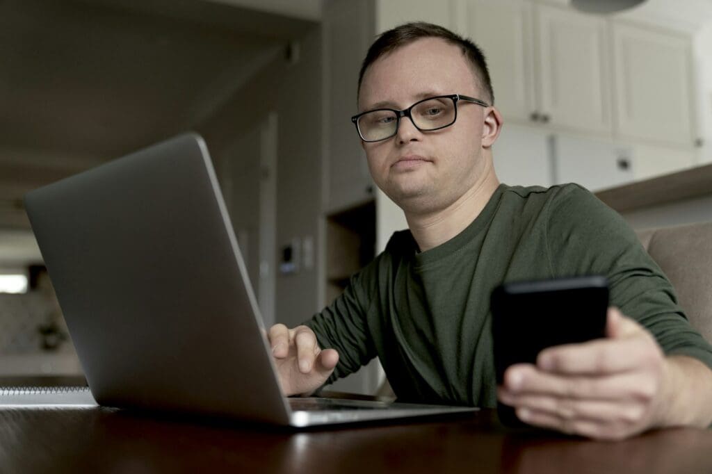 Low angle of adult caucasian man with down syndrome using laptop and phone at home